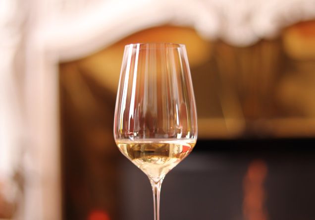 A glass of golden Burgundy Chardonnay during a private at-home tasting session, with a warm blurred fireplace in the background.